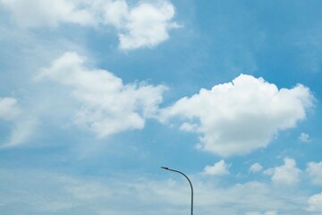 A solitary street lamp curves against a bright blue sky with scattered white clouds, creating a minimalistic and serene urban scene.