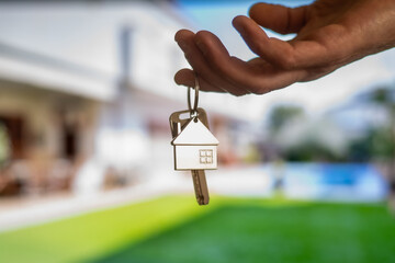 Real estate agent holding house keys with house shaped keychain in front of a blurred background representing a new property with garden and pool