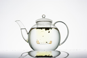 Teekanne aus Glas vor hellem Hintergrund mit Deckel, gefüllt mit Wasser, im Teeeinsatz schwimmt loser Kräutertee, beginnt zu ziehen, Silhouette, minimalistisch