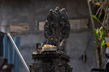 A Balinese stone shrine with intricate carvings, holding a Canang sari offering with flowers and...