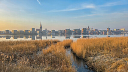 Fototapeta premium scenic view of the city skyline of Rostock, germany, reflecting on a calm river at sunset. A small waterway winds through golden reeds in the foreground