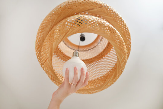 Hand installing white light bulb into a woven rattan lampshade. Close-up shot of home lighting fixture maintenance.