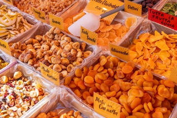 Various dried fruits including figs, apricots, pineapples and pears are displayed in plastic-lined trays with labeled signs at an open air market