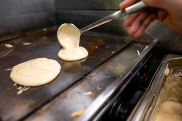 A close-up shot of a hand using a ladle to pour pancake batter onto a hot griddle, with a portion of the batter already sizzling on the surface.