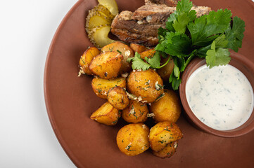 Delicious grilled pork ribs with golden roasted potatoes, pickles, and fresh parsley on a rustic clay plate. Served with creamy white sauce. Perfect homemade meal