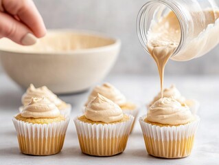A hand is pouring frosting from a jar onto freshly baked cupcakes, set against a bowl in a bright kitchen environment.