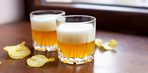 Two glasses of fresh delicious light unfiltered beer and potato chips on wooden windowsill close-up