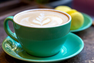 Fresh delicious coffee latte with pattern and yellow macaroon in green dish on table. Selective focus