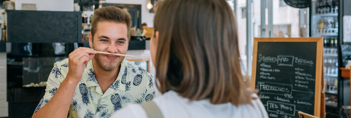 Obraz premium Banner of young man playing with chopstick like it is mustache while enjoying meal with his girlfriend at cafe, sharing laughter and love over their lunch date. Happy couple having fun in restaurant.
