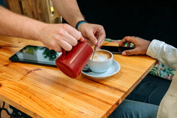 Unrecognizable barista preparing a frothy cappuccino, skillfully pouring creamy milk into a coffee cup on a rustic wooden table in a cozy cafe, while a customer waits nearby, engaged with a tablet