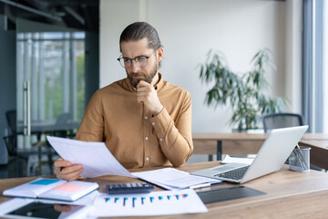 Bearded businessman in glasses examines documents at his desk with a laptop, calculator, and charts, thoughtfully analyzing information.
