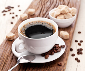 Cup of coffee on white plate, several coffee beans and brown sugar on wooden table close up. Top view.