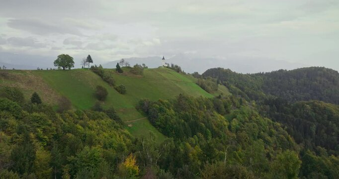 Church of Saints Primus and Felician in the Jamnik region, magnificent view surrounded by lush green hills and mountains. The drone flies over the church and opens up to the wide plain.
