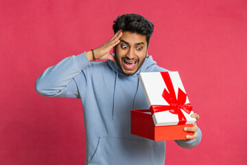 Excited Indian man receiving a festive gift with a red bow opening the box with joy celebrating a...