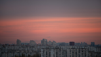 A vibrant sunset casts shades of pink and orange over a city skyline. Tall buildings rise above a cluster of residential apartments, creating a striking urban landscape