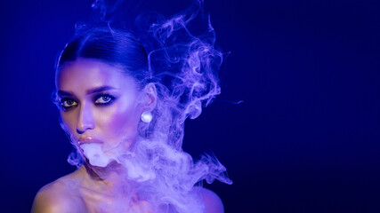 Fototapeta premium Woman vaping and blowing smoke in the air, posing in studio illuminated by blue and pink lights, panorama with free space
