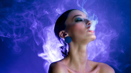 Naklejka premium Woman blowing smoke swirls around her face, posing on studio background illuminated by blue and purple light, panorama