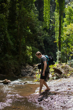 A young male gingerly hikes barefoot across a slippery rainforest stream.