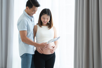 Cheerful Asian pregnant woman or wife with caring husband standing near window at home, smiling and looking at ultrasound image of their baby. Parenthood, bonding and expectant parents