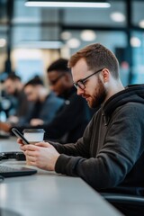 Obraz premium A young adult male is checking his cellphone while sitting alone at a desk in a modern, open office. His focus on his phone suggests multitasking and distractions from work.
