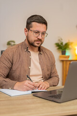Naklejka premium Young businessman writing down or taking down notes while attending online video class or office meeting on laptop computer at home office desk. Guy freelancer support services looking at webcam.