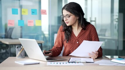 Busy thoughtful young businesswoman analyzing financial documents checking data on laptop sitting at desk at workplace in business office. Financier deals with financial reports, engaged in accounting - Powered by Adobe