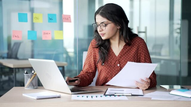 Busy thoughtful young businesswoman analyzing financial documents checking data on laptop sitting at desk at workplace in business office. Financier deals with financial reports, engaged in accounting
