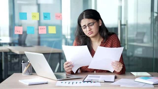 Confused young businesswoman having difficulty with paper work sitting at workplace in business office. Puzzled female employee looks at documents and cannot understand problem and looking at camera