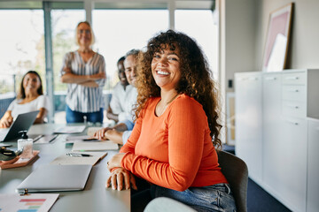 Confident woman smiling in a modern office meeting setting. She looks directly into the camera, smiling, while her happy diverse colleagues sit behind her.