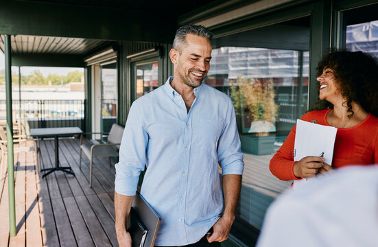 Two business professionals sharing A laugh on modern office balcony. They are taking a break from a meeting, standing with their laptops and notebooks.