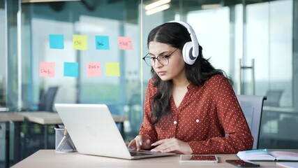 Confident young businesswoman in wireless headphones typing on laptop at workplace in a modern business office. Focused female IT specialist in glasses works on computer while listening to podcast