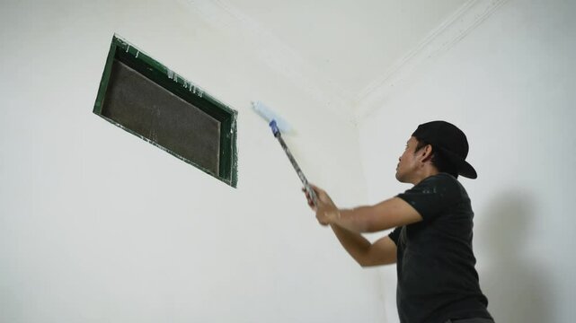 Asian man repairman painting walls of apartment in white using paint roller, 4k video. House finishing services concept.