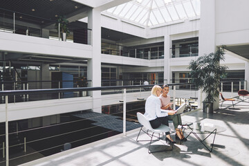 Diverse business women discussing paperwork together. They are sitting in a hallway, in a large open minimalist concrete community
