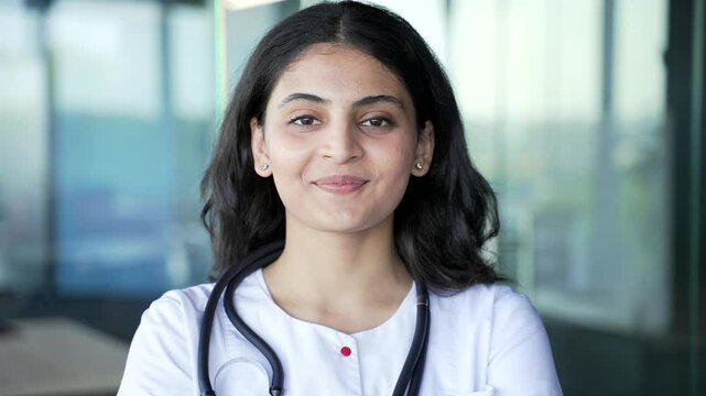 Portrait of young smiling female doctor in white coat looking at camera in hospital clinic. Headshot of happy woman medical worker physician poses standing at workplace in modern office. Close up
