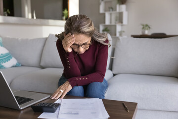 Older Hispanic woman sits at table with bills and laptop with stressful expression, experiences...