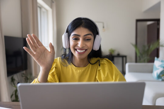 Cheerful Indian woman wearing headphones waving at screen seated at desk, engaged in video conferencing on her laptop, start personal or business digital communication through video call application