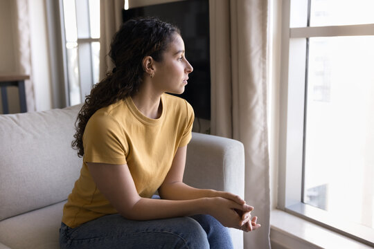 Young Brazilian woman sitting on sofa, gazing out of window with pensive and contemplative expression, deep in thought, looking concerned, feeling longing, reflects on personal matters or loneliness