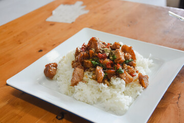 square white plate containing topped with a stir-fried dish. The stir-fried portion includes pieces of meat, likely pork, accompanied by vegetables like red bell peppers and fresh green herbs.
