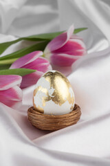 Golden Easter egg in a nest with pink tulip flowers close up on a white silky backgorund, Easter banner.