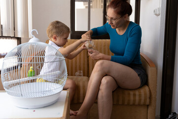 Mother and son feeding a pet budgie in a cage at home