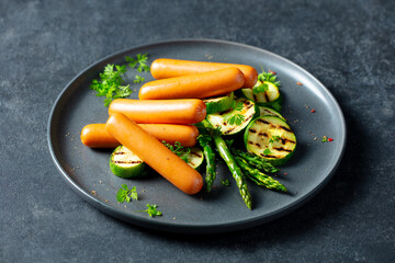 Sausages with grilled zucchini, asparagus and fresh herbs on a plate. Dark background. Close up.