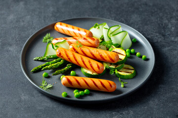 Grilled sausages with asparagus, zucchini, green peas and cucumber salad on a plate. Dark grey background. Close up.