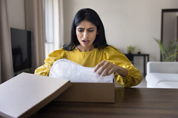 Indian woman sits at table unboxing package with dissatisfied and shocked expression, received...