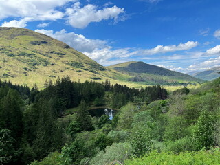 View of mountain and forest landscape in Glencoe, Scotland. Green hills, pine trees, calm lake and a scenic valley under a blue sky with clouds.