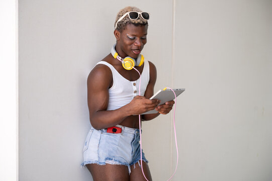 Stylish young african american non-binary person using digital tablet while wearing headphones and sunglasses