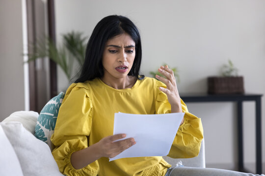 Upset Indian woman sit on sofa holding letter or document, received unexpected bill or invoice, job, university or application rejection letter, unexpected deduction in bank account, fine or penalty