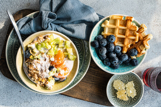 Overhead view of a Breakfast selection of fresh waffles with blueberries, yogurt with granola, kiwi fruit, tangerines, grapes, honey and a glass of fresh fruit juice