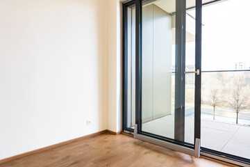 Modern tilt and slide glass door system for balcony access in a residential apartment. Minimalist design with aluminum frame, wooden flooring, and natural daylight. Indoor perspective.