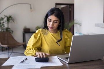 Focused Indian woman sitting at desk with laptop, using calculator, calculates expenses, managing...