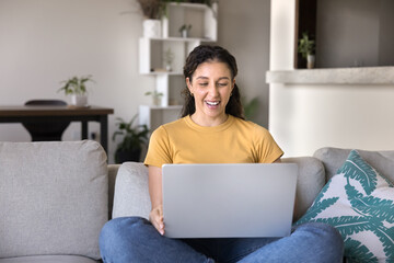 Naklejka premium Young Latina woman laughing while watching online movie via subscription streaming services, resting on couch with modern laptop, enjoy videos and on-line content spend carefree pastime alone at home